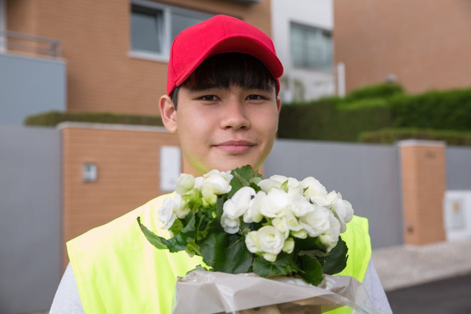 A young delivery person with light skin, wearing a red cap and neon yellow safety vest, is holding a bouquet of fresh white roses with green leaves. The bouquet is wrapped in white paper, and the pers
