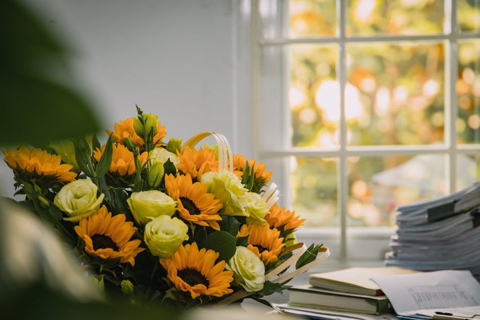 A vibrant bouquet of fresh flowers resting on a desk near a large window bathed in natural light. The arrangement features bright yellow sunflowers with dark brown centers, creamy white roses, and gre