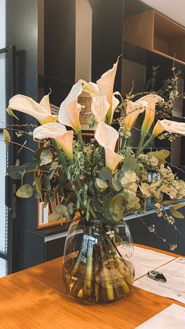 A clear glass vase filled with a fresh floral arrangement featuring large, creamy-white calla lilies with smooth, trumpet-shaped petals, accented by small white gypsophila clusters and green eucalyptu