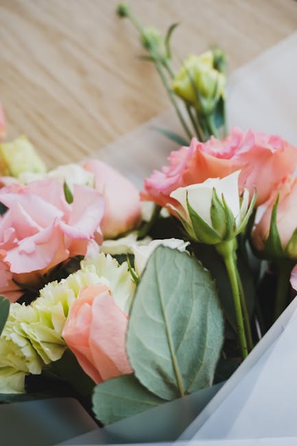 A close-up of a floral arrangement featuring soft pink and cream eustoma (lisianthus) flowers with delicate, ruffled petals, interspersed with fresh green foliage and large, smooth leaves. The bouquet